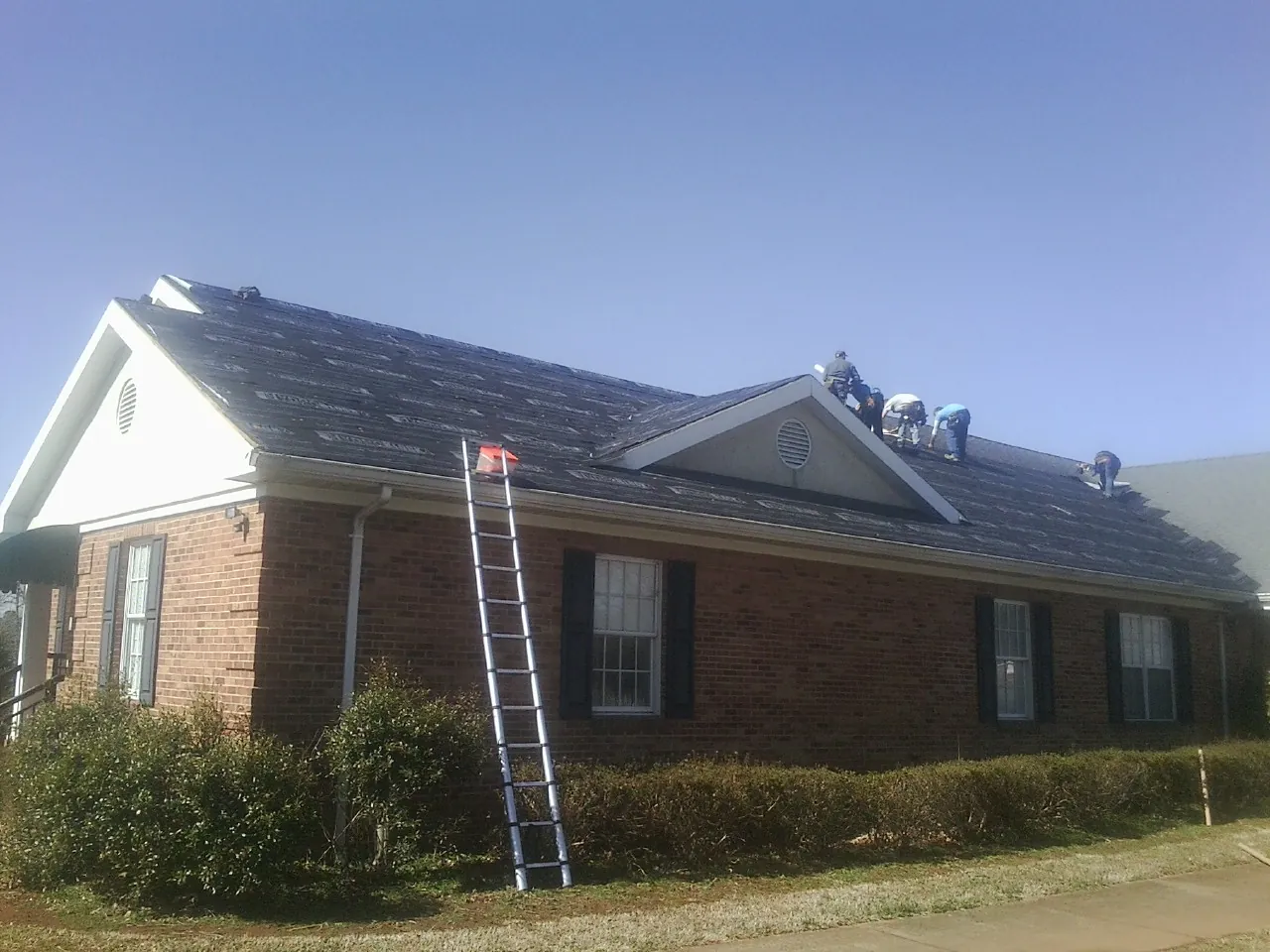 Crew tearing shingles off a brick ranch roof with a ladder set — Metal Roof Repair work by our crew in Spencer